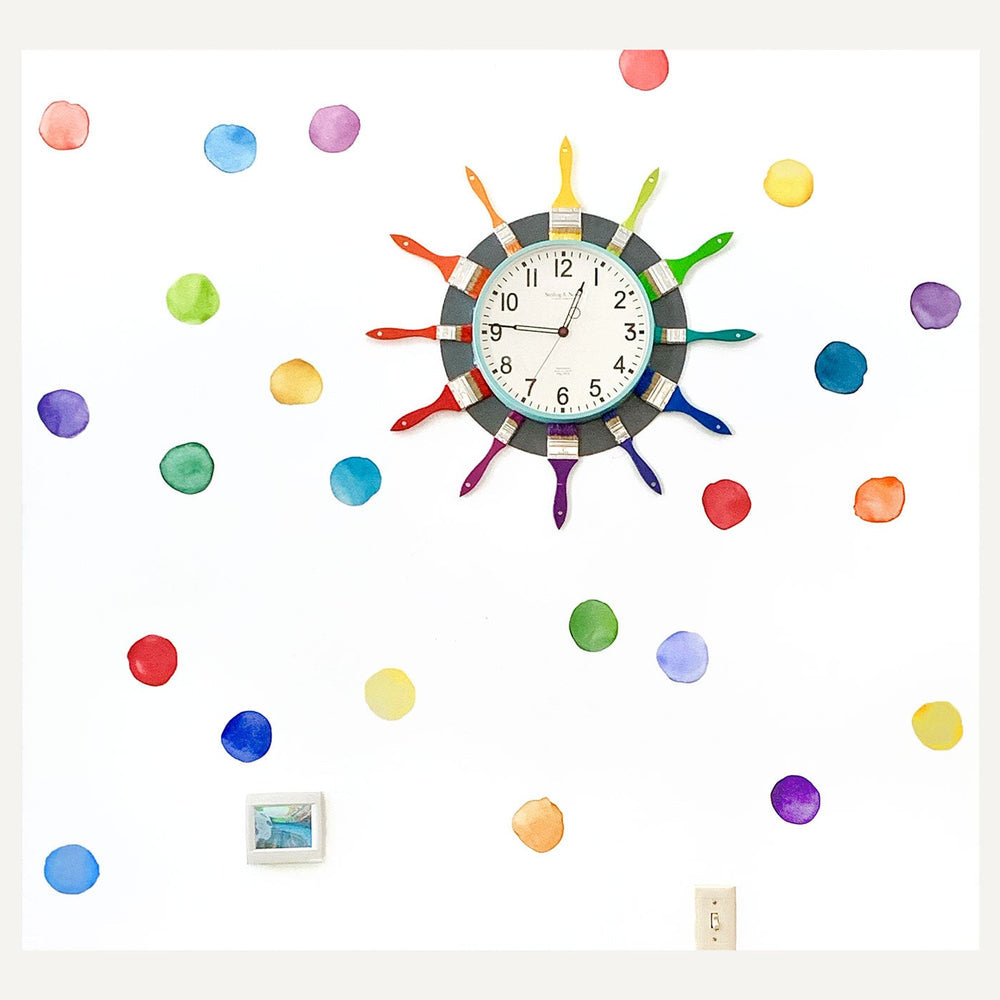 Art classrooom wall is adorned with watercolor dots and features a clock decorated with rainbow paintbrushes. 