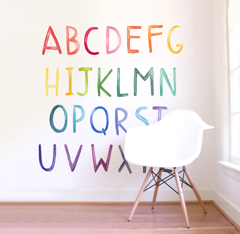 Watercolor uppercase alphabet fabric decals in rainbow hues arranged on a white nursery wall with modern wooden chair
