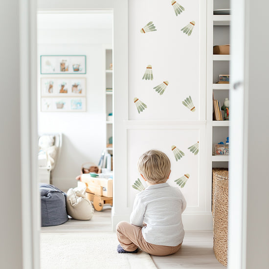 Watercolor badminton birdies decals by Mej Mej are tossed on an accent wall in a playroom as if they there has just been a strike. A little boy is sitting on the floor looking at them.