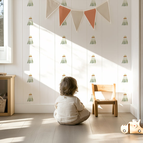 Child sitting on floor in sunlit kids’ room with Mej Mej watercolor fabric wall decals of badminton birdies in a grid pattern on the wall with color-coordinated pennant hanging in front.