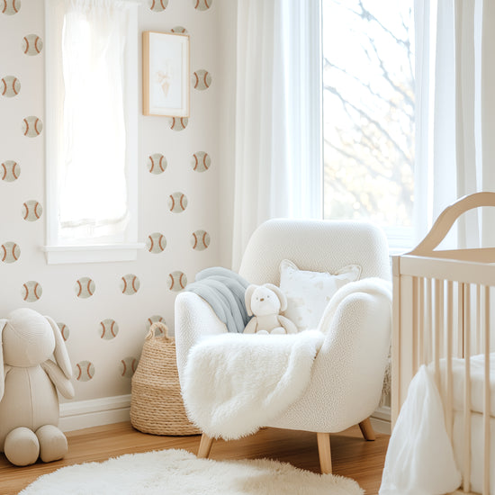 A wall decorated with watercolor baseball wall decals by Mej Mej is next to a window in a child’s nursery of soft muted creams with a natural crib and soft cream chair.