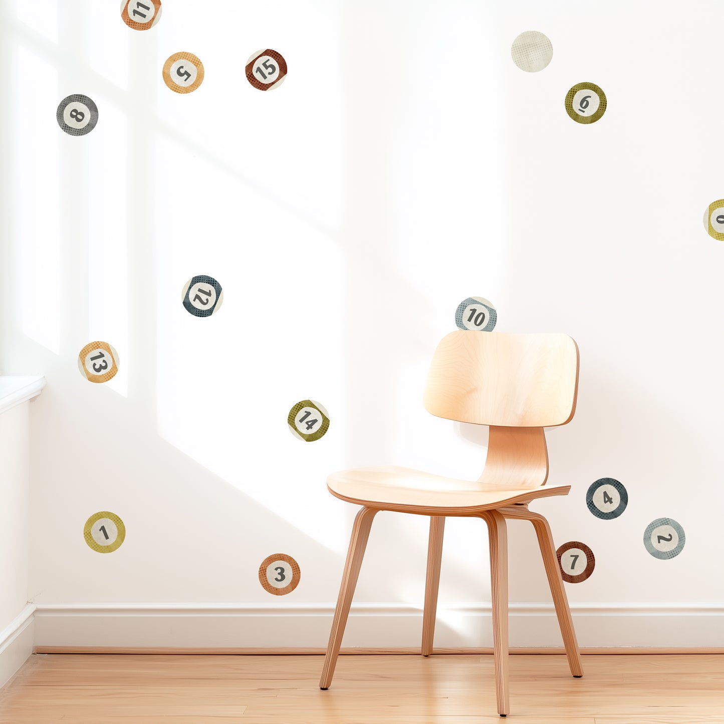 A wall is decorated with watercolor billiard ball wall decals by Mej Mej. They are placed in a loose toss pattern, as though someone has taken the first shot of the game. A modern chair sits in front of them for scale.