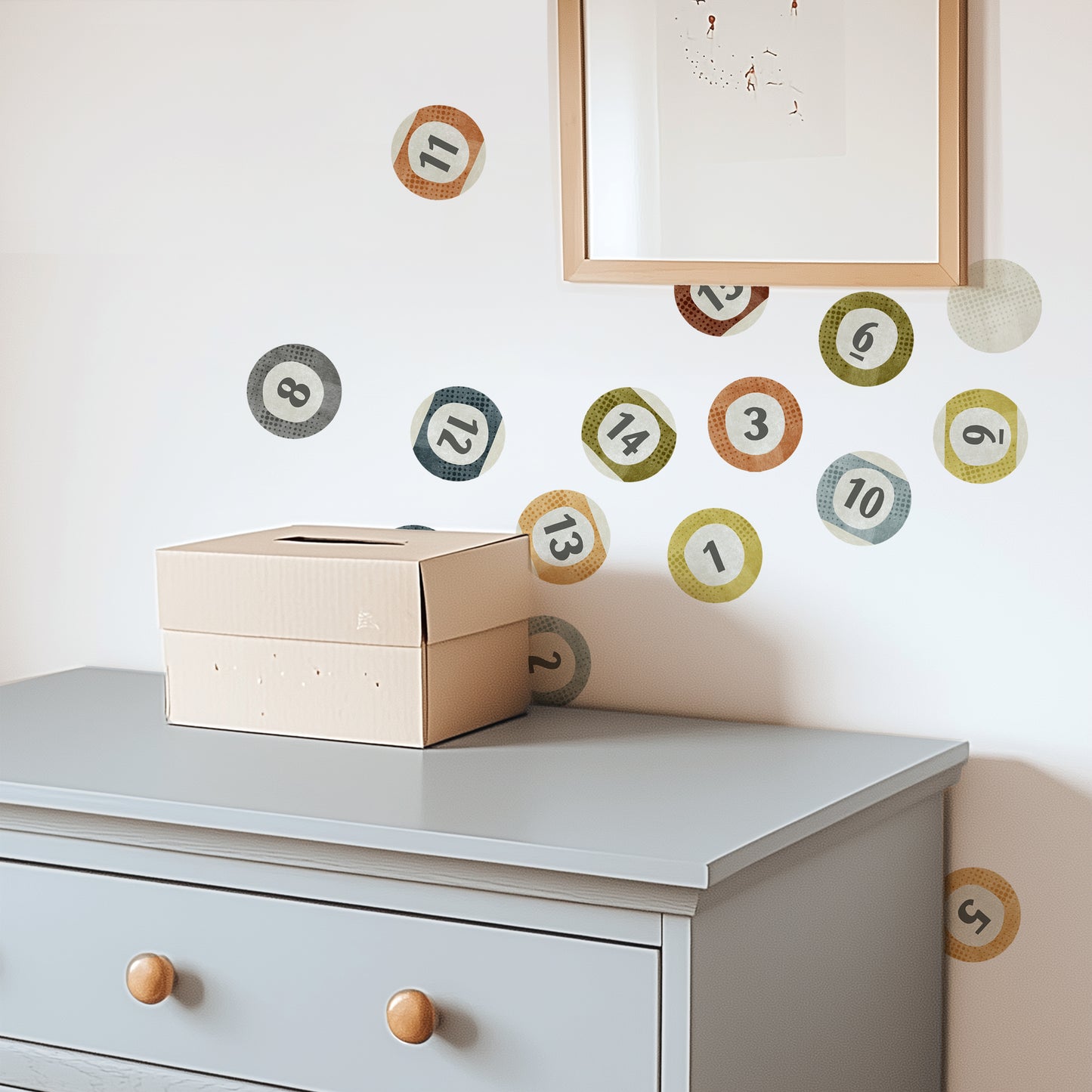 A bedroom wall is decorated with watercolor fabric wall decals featuring billiard balls by Mej Mej. The balls are painted in rich but muted colors. A chest of drawers sits in front of the wall and a framed piece of art hangs on top of the decals.