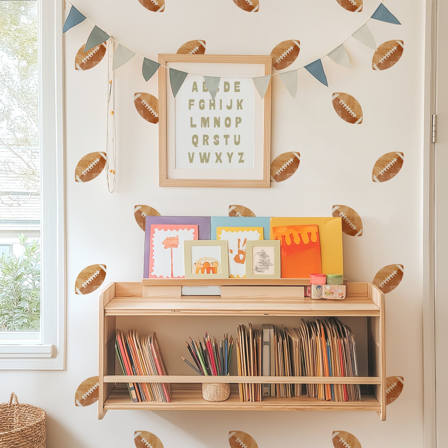 Watercolor football wall decals adorn a wall behind a bookshelf with books, art supplies and kids’ artwork displayed. A framed alphabet and pennant are hung on top of decals.