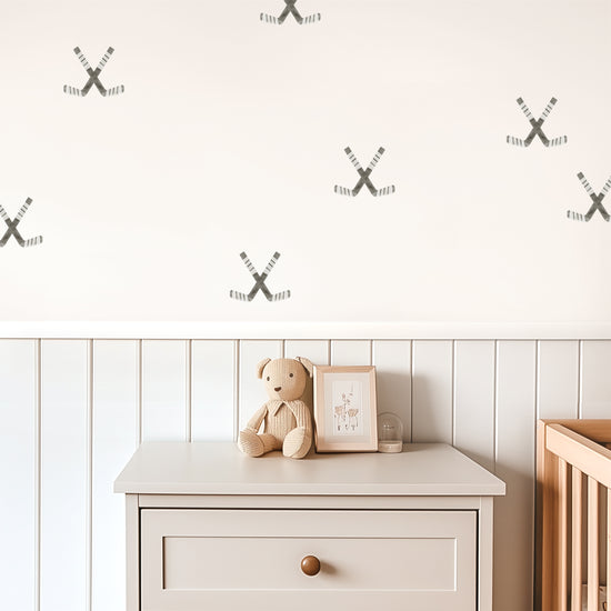 Watercolor crossed hockey sticks removable decals by Mej Mej are tossed in a directional pattern on a wall in a child’s playroom. A chest and crib sit in front of a chair railing.