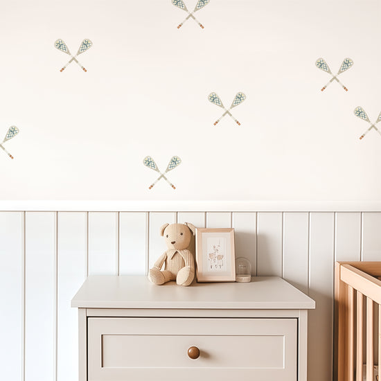 Watercolor crossed lacrosse sticks removable decals by Mej Mej are tossed in a directional pattern on a wall in a child’s playroom. A cream side table and crib sit in front of a chair railing.