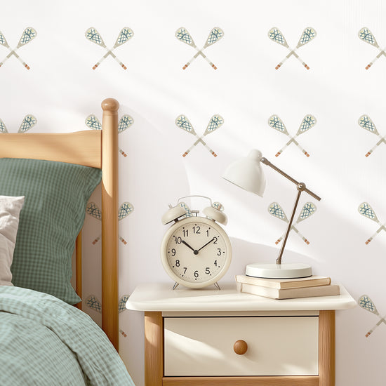 A white wall with crossed lacrosse sticks fabric decals placed in a grid pattern is the backdrop for a stylish bedroom. A bed with coordinating bedding, a nightstand with an oversized vintage alarm clock and gooseneck lamp complete the modern vibe.