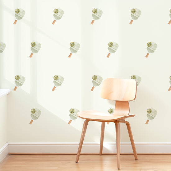 A wall is decorated with removable fabric wall decals of pickleball paddles with balls by Mej Mej. They are placed in a modern grid pattern. A mid-century chair sits in front of them for scale.