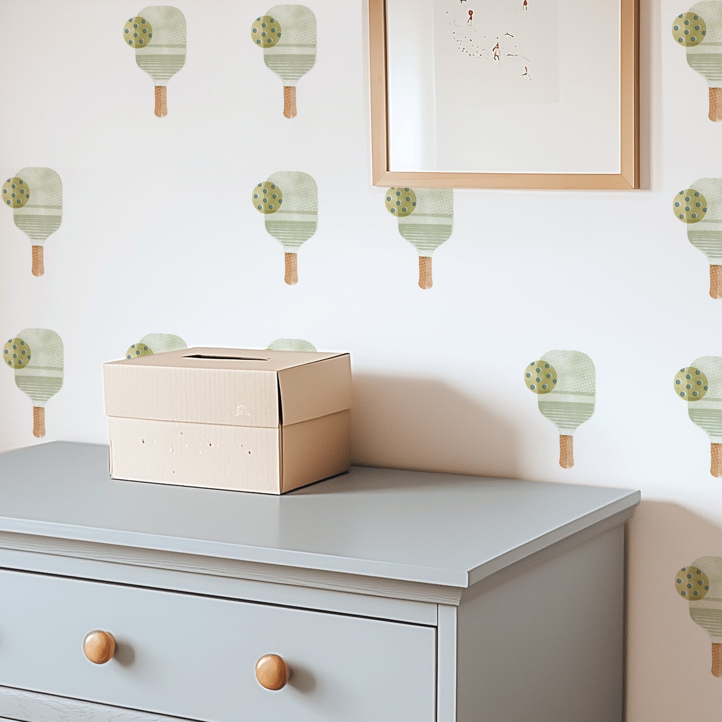 A bedroom wall is decorated with watercolor fabric wall decals featuring pickleball paddles with balls by Mej Mej. A chest of drawers sits in front of the wall and a framed piece of art hangs on top of the decals.