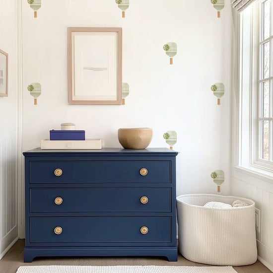 Watercolor pickleball paddles with balls removable decals by Mej Mej are placed in an open grid on an accent wall in a dressing area behind a navy chest of drawers and basket of blankets.