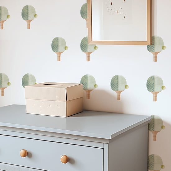 A bedroom wall is decorated with watercolor fabric wall decals featuring ping pong paddles with balls by Mej Mej. A chest of drawers sits in front of the wall and a framed piece of art hangs on top of the decals.