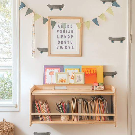 Watercolor skateboards fabric wall decals adorn a wall behind a bookshelf with books, art supplies and kids’ artwork displayed.