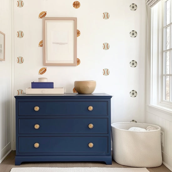 Watercolor Sports Balls Wall Decals by Mej Mej are placed in an open grid on an accent wall in a dressing area behind a navy chest of drawers and basket of blankets.