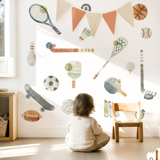 Child sitting on floor in sunlit kids’ room with Mej Mej watercolor fabric wall decals of sports equipment in a grid pattern on the wall with color-coordinated pennant hanging in front.