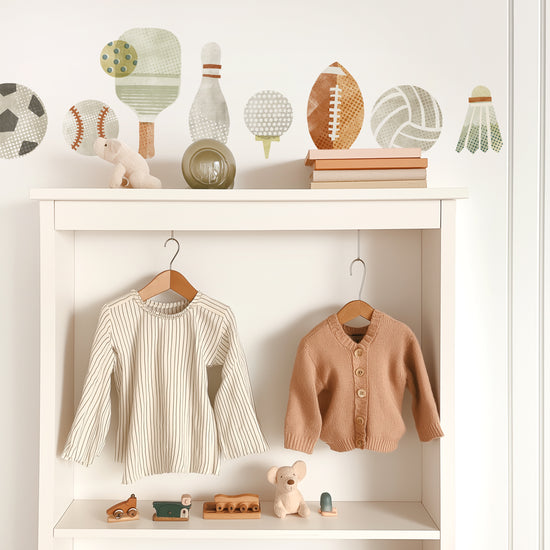 Removable fabric wall decals by Mej Mej are placed in a horizontal line above a contemporary white open-backed shelf unit with cute children’s clothes hanging on wooden hangers.