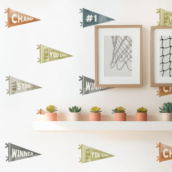 An even grid of watercolor Sports Pennants Wall Decals by Mej Mej adorns a wall in a sunny room. A hanging narrow shelf displays succulents.