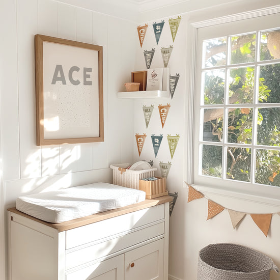 Watercolor Sports Pennants Wall Decals by Mej Mej are placed in a grid pattern on an accent wall in a sunlit child’s nursery. A changing table is in front of the accent wall with a framed print that says ACE.