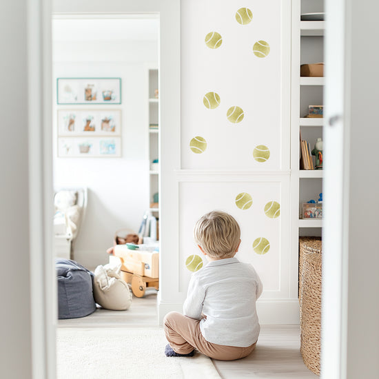 Watercolor tennis balls decals by Mej Mej are tossed on an accent wall in a playroom. A little boy is sitting on the floor looking at them.